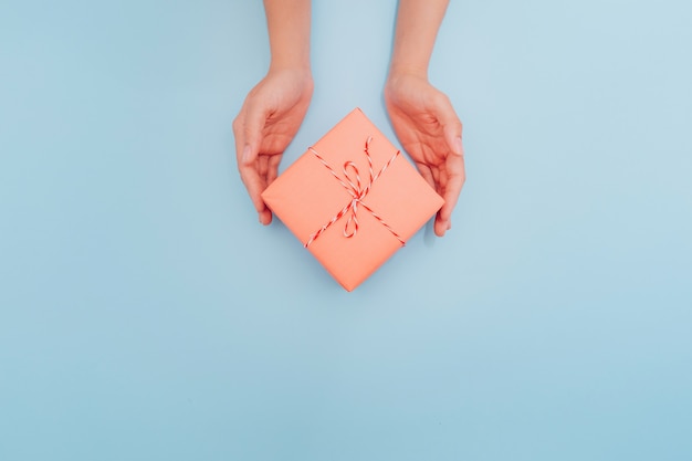 Mãos femininas estão segurando uma caixa de presente rosa com uma fita fina como um presente para o natal, ano novo, dia das mães ou aniversário em um fundo azul tabela, vista superior. lugar para texto.