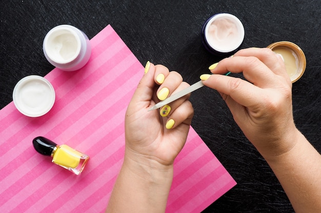 Mãos femininas fazem manicure. na mesa preta há potes de creme, verniz e um guardanapo rosa.