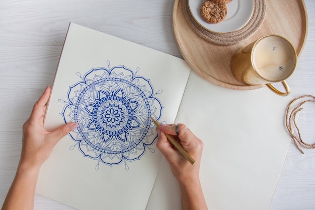 Mãos femininas fecham desenhar mandala floral redonda decorativa. hobby e relaxamento em casa. uma caneca de café e biscoitos em uma bandeja de madeira. fundo branco.