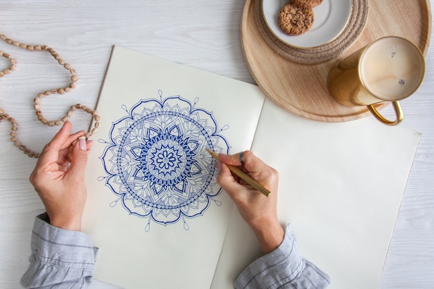 Mãos femininas fecham desenhar Mandala floral redonda decorativa. Hobby e relaxamento em casa. Uma caneca de café e biscoitos em uma bandeja de madeira. Fundo branco.