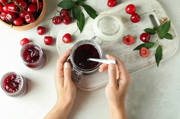 Mãos femininas, geléia de cereja e ingredientes na mesa texturizada branca