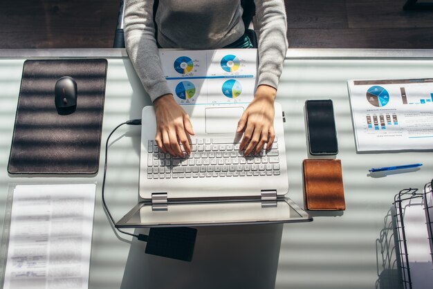 Mãos femininas na mesa de trabalho com laptop e documentos vista de cima.