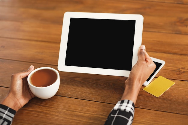 Mãos femininas negras segurando um tablet digital com tela em branco para propaganda. Garota afro-americana, bebendo café, usando smartphone e cartão de crédito em um fundo de mesa de madeira.