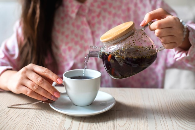 Mãos femininas seguram bule transparente com chá de ervas e despejam chá quente em xícara branca