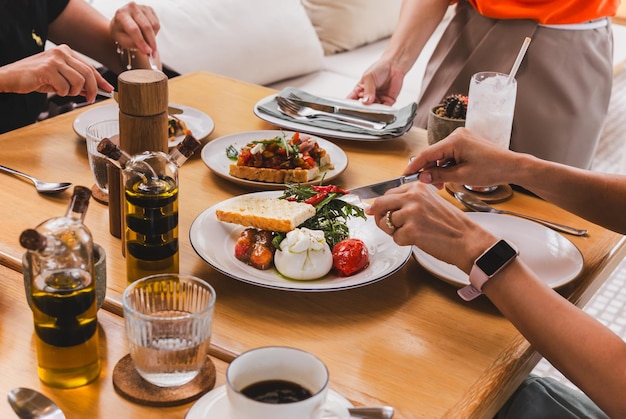 Mãos femininas seguram faca e garfo cortando salada de foguete com queijo burrata italiano e tomate em prato branco