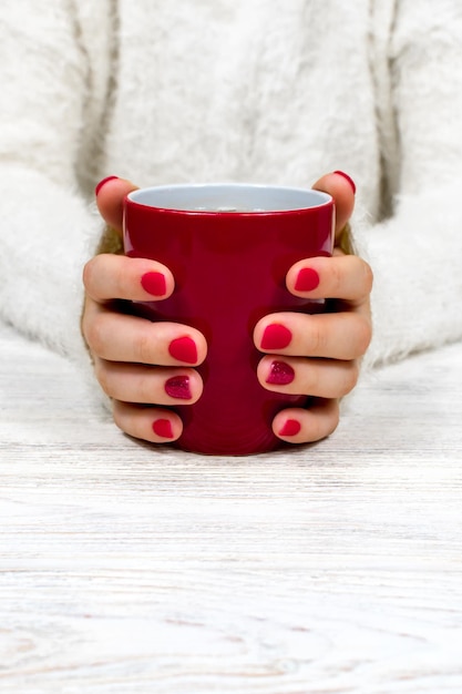 Foto mãos femininas seguram um copo vermelho contra o fundo de um suéter fofo branco com uma manicure