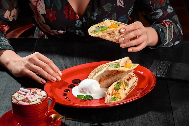 Mãos femininas segurando rolo de frutas em lavash fino servido com sorvete e xícara de cacau