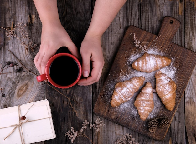 mãos femininas segurando um copo de cerâmica vermelha com café preto ao lado de croissants assados em pó com açúcar em pó