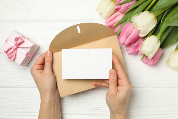 Mãos femininas segurando um envelope com um cartão em branco no fundo branco de madeira com tulipas e uma caixa de presente