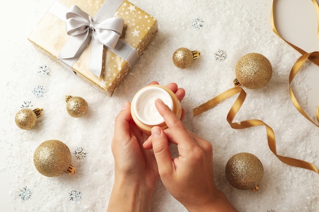 Mãos femininas segurando um frasco de creme cosmético no fundo com acessórios de natal