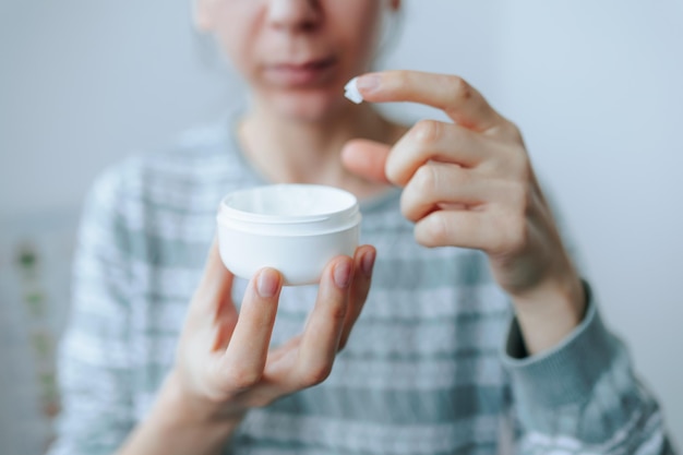 Mãos femininas segurando um pote de creme facial esfregando hidratante aplicando creme na pele do rosto