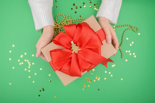 Mãos femininas segurando um presente de Natal com laço vermelho em verde. Postura plana