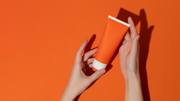 Mãos femininas segurando um tubo cosmético de plástico laranja em branco na maquete de fundo laranja
