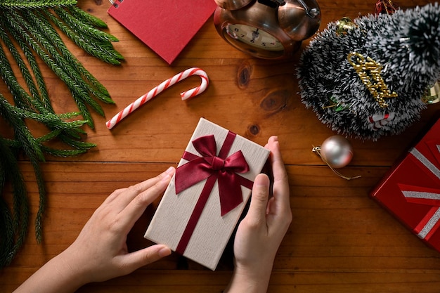 Mãos femininas segurando uma caixa surpresa de presente de Natal sobre mesa de madeira com decoração de Natal