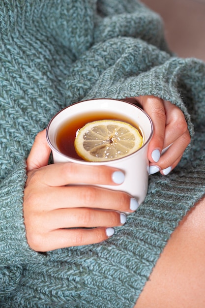 Mãos femininas segurando uma caneca de chá quente com limão de manhã. Mulher jovem relaxante xícara de chá nas mãos.