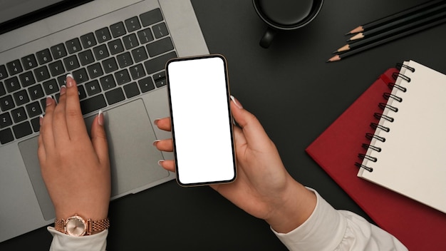 Mãos femininas segurando uma maquete de smartphone e digitando trabalho no laptop na mesa de escritório preta moderna