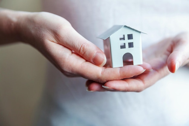 Mãos femininas segurando uma pequena casa de brinquedo branco em miniatura Sonho de seguro de propriedade hipotecária movendo-se para casa e conceito imobiliário