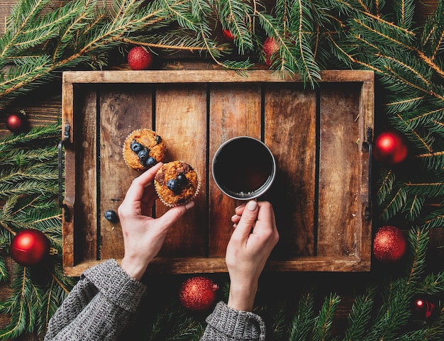 Mãos femininas segurando uma xícara de chá ao lado da árvore de Natal e muffins em uma mesa