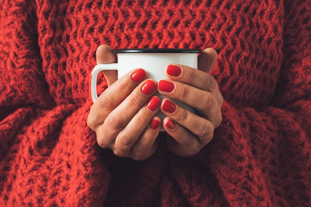 Mãos femininas segurando uma xícara de chá de metal branco contra um suéter de inverno de malha vermelha Conceito mínimo