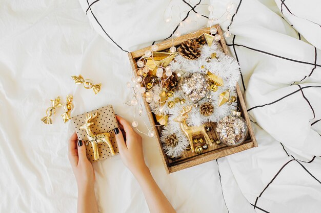 Mãos femininas segurar um presente. Conjunto de decorações de Natal em caixa de madeira na cama branca com um cobertor. Conceito de férias. Camada plana, vista superior