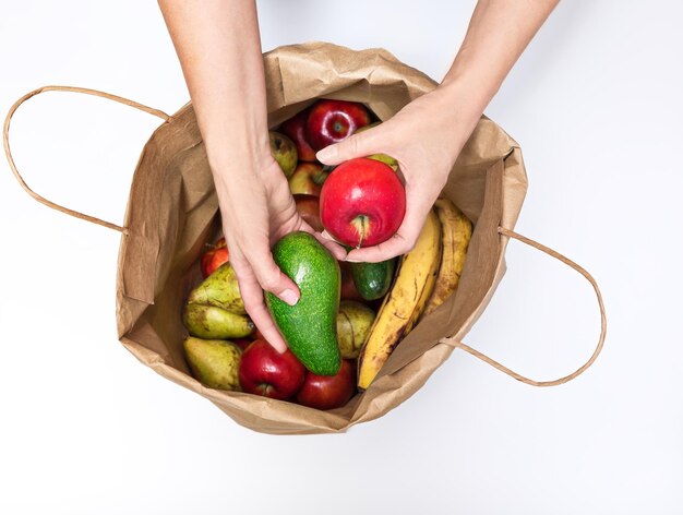 Mãos femininas tiram uma maçã vermelha e um abacate de um saco de papel cheio de diferentes frutas isoladas em um fundo branco Entrega e compra de alimentos ecológicos saudáveis