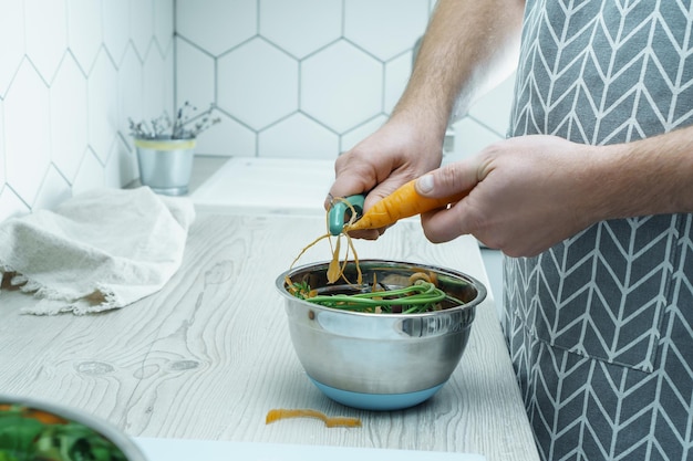 Mãos masculinas descascando cenoura fresca com descascador sobre tigela de cascas de vegetais Restos de comida Coleta separada de lixo