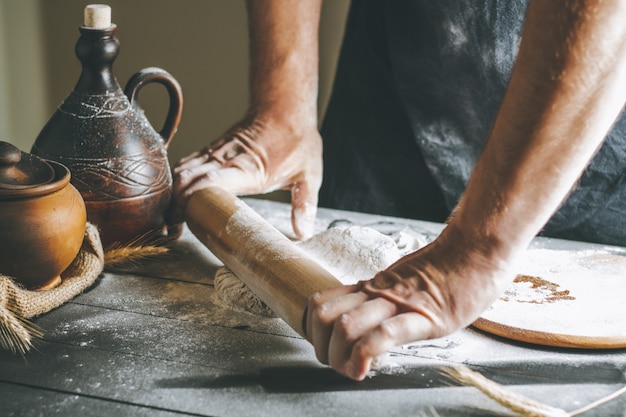 Mãos masculinas enrolam massa e farinha com o rolo ao lado da panela de barro e uma garrafa de óleo na mesa escura, enquanto cozinham