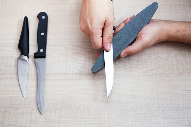 Foto mãos masculinas fortes de close-up afiam uma faca de metal de cozinha com uma pedra de amolar. afiação de facas para uso doméstico. vista do topo. grupo de facas
