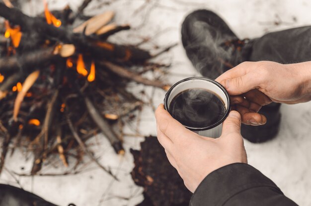 Mãos masculinas seguram uma caneca de café perto de uma fogueira em chamas.