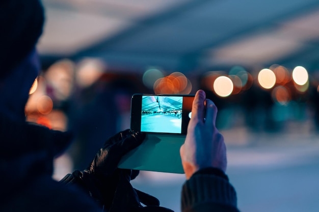 Mãos masculinas segurando um telefone celular no modo de câmera fotográfica - homem tirando foto de uma pista de patinação no gelo à noite