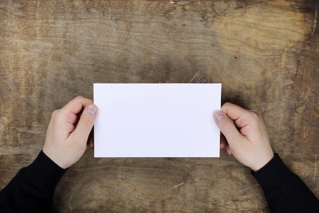 Mãos masculinas segurando uma folha de papel em branco no fundo de uma mesa de textura de madeira