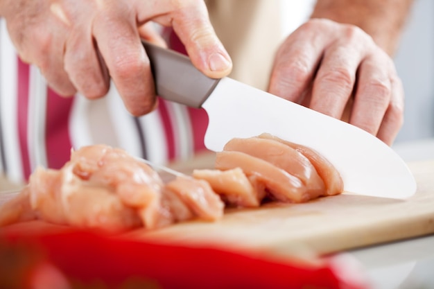 Mãos masculinas sênior cortando filé de frango na placa da cozinha.