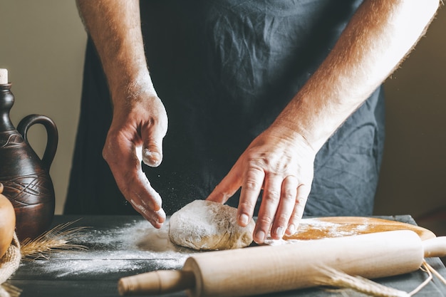 Mãos masculinas sove a massa com farinha ao lado de uma panela de barro, uma garrafa de óleo e um rolo na mesa escura, enquanto cozinha