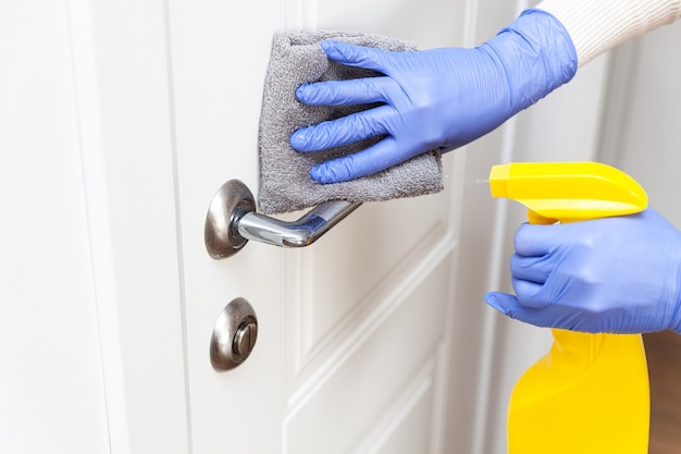 Mãos nas luvas, desinfetando a maçaneta da porta com pano e detergente em spray