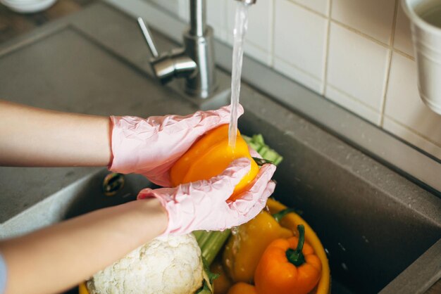 Mãos nas luvas lavando pimenta sob o fluxo de água na pia durante a epidemia de vírus Lavando legumes Mulher com mãos cor de rosa limpando legumes frescos preparando-se para cozinhar a refeição na cozinha moderna