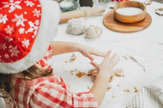 Mãos parcialmente desfocadas de uma menina com boné de Natal e camisa quadriculada tricotar massa para biscoitos na mesa branca