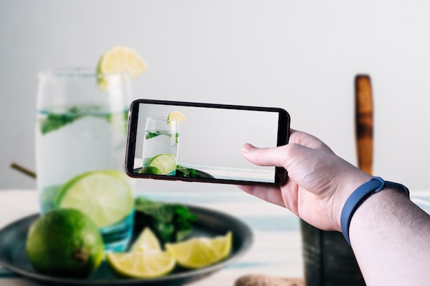 Mãos pegando um smartphone tirando uma foto de um mojito com limões em uma mesa de madeira. Espaço para texto. Foco seletivo