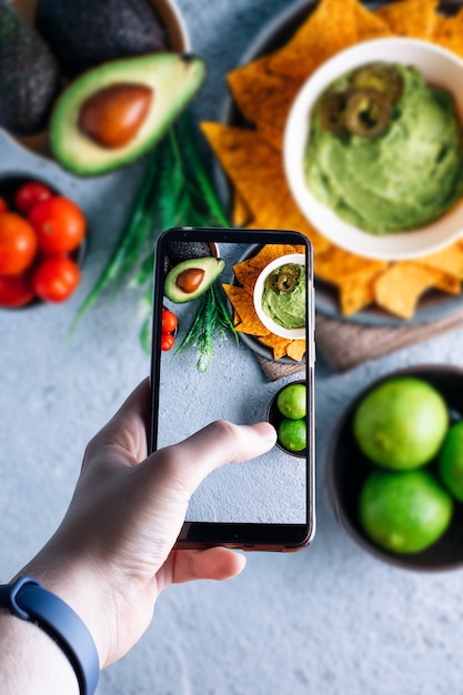 Foto mãos pegando um smartphone tirando uma foto de uma tigela com guacamole. espaço para texto. foco seletivo