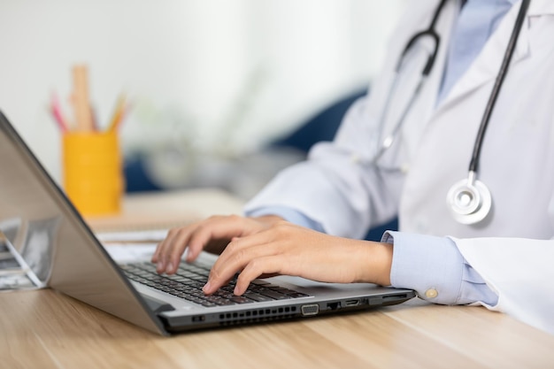 Foto mãos próximas de um médico digitando em um computador portátil na mesa doutor com estetoscópio trabalhando em um consultório médico mãos digitando no teclado para gravar o histórico médico do paciente no hospital