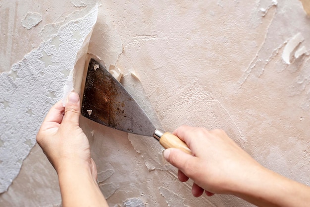 Mãos removendo papel de parede antigo com a ajuda de uma espátula durante o reparo na sala