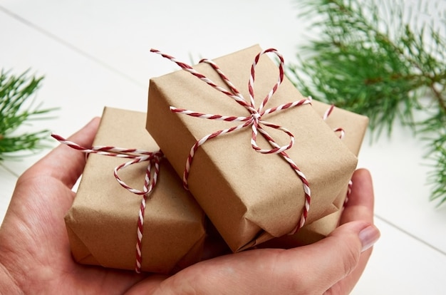 Mãos segurando caixas de presente de Natal sobre uma mesa branca com galhos de pinheiros
