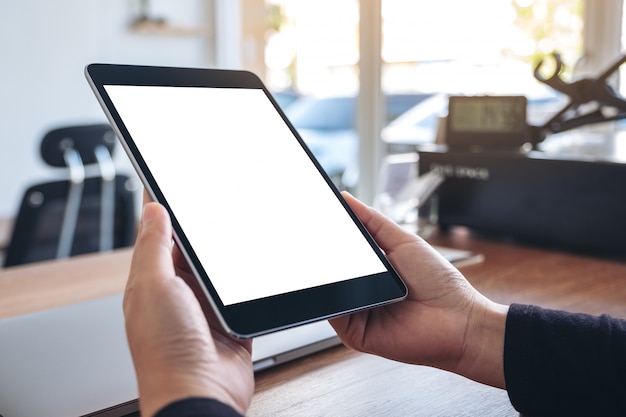 mãos segurando e usando tablet pc preto com tela de desktop em branco branco com notebook na mesa de madeira no escritório