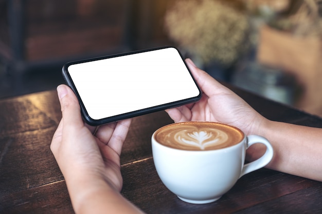 mãos segurando e usando um telefone celular preto com tela em branco horizontalmente para assistir com uma xícara de café na mesa de madeira