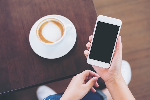 Mãos segurando o telefone celular branco com tela preta em branco com copo de café na mesa de madeira