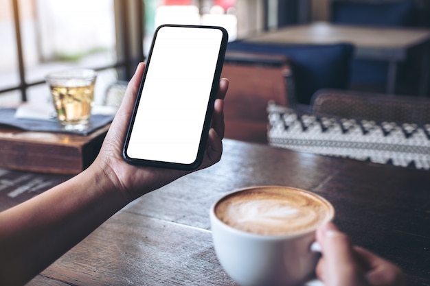 Mãos segurando um celular preto com tela em branco enquanto bebe café em um café vintage