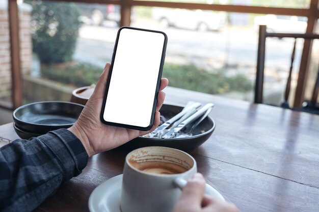 mãos segurando um celular preto com tela em branco enquanto bebe café no café