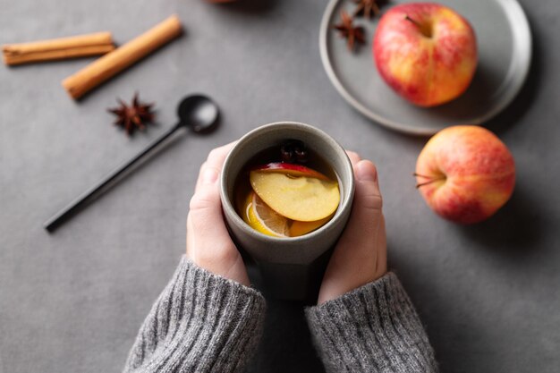 Mãos segurando um copo cinza com ponche de maçã caseiro ou sidra com maçãs e canela em um fundo escuro com frutas frescas e especiarias