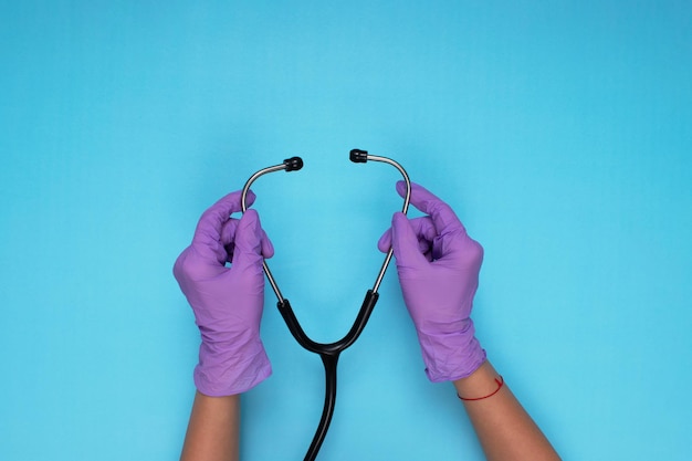 Mãos segurando um estetoscópio com luvas médicas de cor lilás sobre fundo azul claro