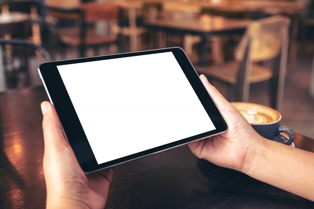 mãos segurando um tablet pc preto com uma tela em branco e uma xícara de café na mesa de madeira no café