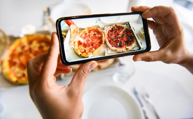 Mãos segurando um telefone celular e tirando a imagem de uma deliciosa pizza recém-assada servida em uma mesa com toalha de mesa branca. Telefone celular em regime de modo ao vivo